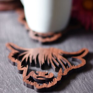 May include: Wooden coaster featuring a Highland cow design. The coaster is brown and has a detailed cutout of the cow's face and horns. A white mug sits on a similar coaster in the background, with a pink flower.