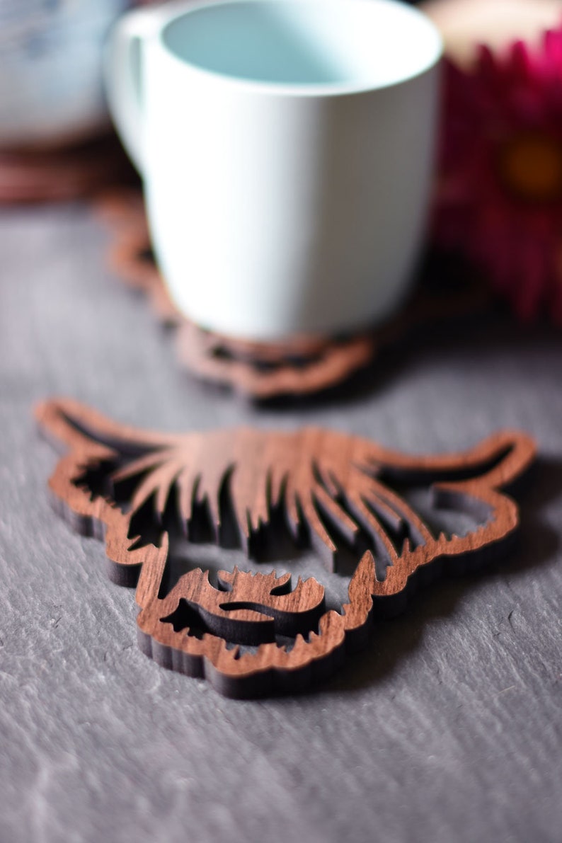 May include: Wooden coaster featuring a Highland cow design. The coaster is brown and has a detailed cutout of the cow's face and horns. A white mug sits on a similar coaster in the background, with a pink flower.