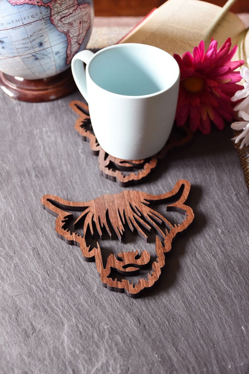 May include: Wooden Highland cow-shaped coasters. One coaster has a light blue mug on top. The coasters are made of dark brown wood and feature a detailed cutout design of a Highland cow's face. The background includes a globe, a book, and flowers.