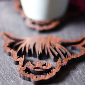 May include: Wooden coaster featuring a Highland cow design. The coaster is brown with a dark outline, and the design is laser-cut. A light blue mug sits on a similar coaster in the background. The coasters are on a gray surface.