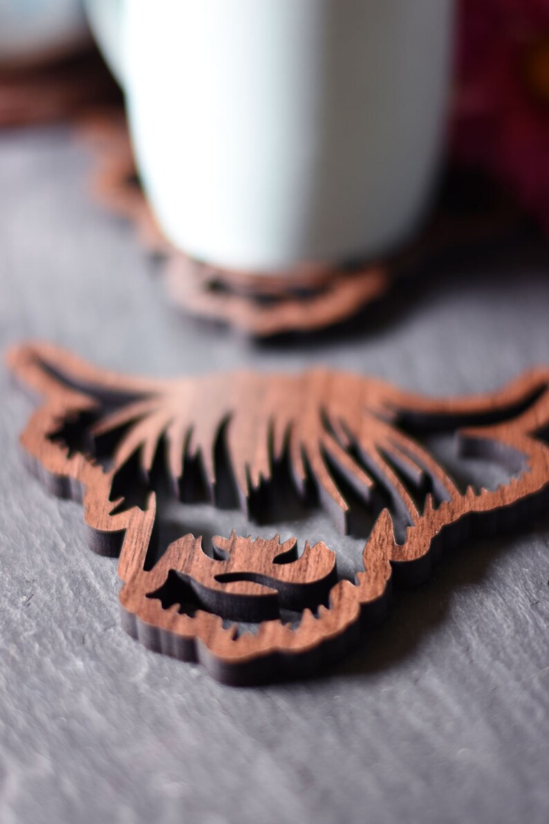 May include: Wooden coaster featuring a Highland cow design. The coaster is brown with a dark outline, and the design is laser-cut. A light blue mug sits on a similar coaster in the background. The coasters are on a gray surface.