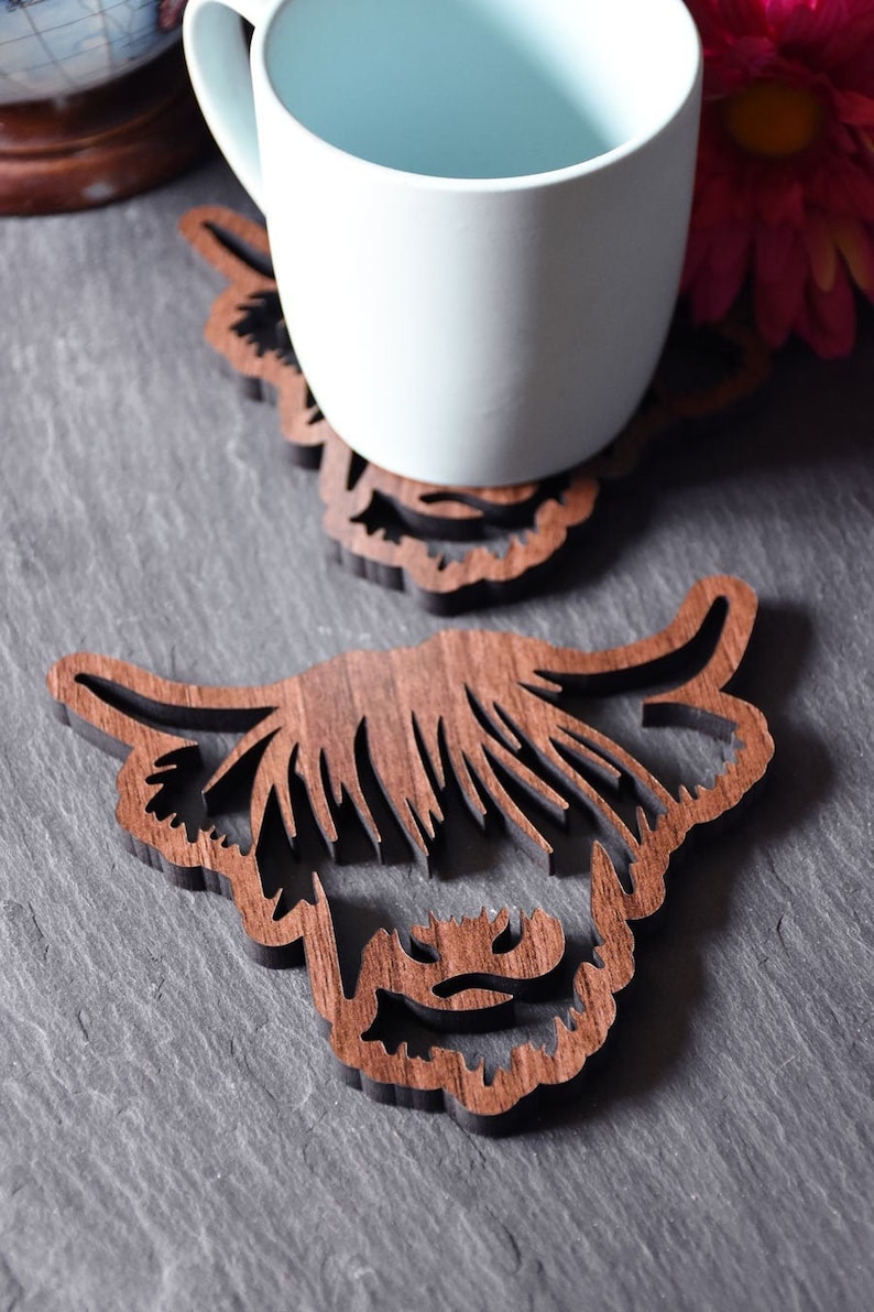 May include: Two wooden coasters with a Highland cow design. One coaster supports a white mug. The coasters are made of dark brown wood and are cut in the shape of a Highland cow's head. The coasters are on a gray surface.