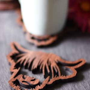 May include: Wooden Highland cow coasters. The coasters are brown and feature a detailed cutout design of a Highland cow's head. A white mug sits on one of the coasters. The coasters are designed to protect surfaces from heat and moisture.
