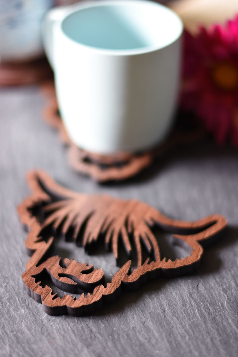 May include: Wooden Highland cow coasters. The coasters are brown and feature a detailed cutout design of a Highland cow's head. A white mug sits on one of the coasters. The coasters are designed to protect surfaces from heat and moisture.