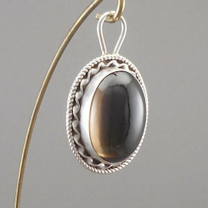 May include: Oval pendant with a polished, dark brown gemstone set in a silver frame. The frame has a rope-like border and a simple bail for a chain. The pendant is displayed on a gold-colored jewelry stand.