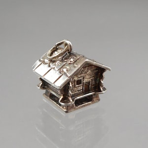 May include: A small, silver-toned charm in the shape of a rustic cabin. The charm features a detailed roof, windows, and decorative elements. It has a loop for attaching to a bracelet or necklace. The cabin charm is a miniature representation of a cozy home.