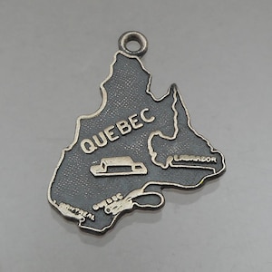 May include: A silver-colored pendant in the shape of the Canadian province of Quebec. The word "QUEBEC" is prominently displayed, along with the names "MONTREAL" and "LABRADOR". A small image of a building is also visible. The pendant has a loop at the top for attaching to a chain.