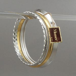 May include: A set of three bangle bracelets in mixed metal tones, featuring one gold-tone band and two silver-tone bands with a twisted rope texture. A small rectangular tag with a dark red background and the gold text Monet N.Y.C. is attached.