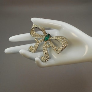 May include: A vintage bow brooch, encrusted with clear rhinestones, and a central emerald-green stone. The brooch is set in a silver-tone metal. The bow design is a classic and elegant accessory.