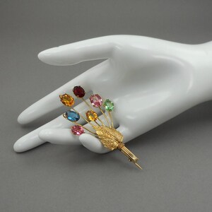 Könnte beinhalten: Eine goldfarbene Brosche in Form eines Blumenstraußes mit farbigen, oval geschliffenen Edelsteinen. Die Brosche hat ein Blatt- und Stieldesign mit Edelsteinen in Rot, Orange, Blau, Rosa, Gelb und Grün. Die Brosche ist etwa 7.6 cm lang.