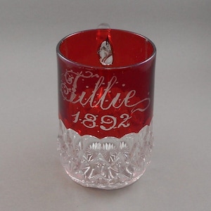 May include: A vintage red glass mug with a clear, patterned base and a red rim. The name "Tillie" and the year "1892" are etched in a cursive font on the red portion of the mug.