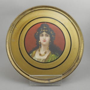 May include: A round, antique portrait of a woman with long dark hair, wearing a gold necklace and earrings. The portrait is set within a gold-toned frame with a red background. The frame has a decorative edge and is supported by a clear stand.