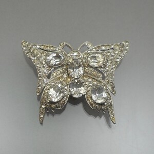 May include: A silver-toned butterfly brooch featuring five large, clear oval-shaped crystals at the centre and wings, surrounded by numerous smaller, pavé-set clear rhinestones covering the entire surface of the metal butterfly wings.
