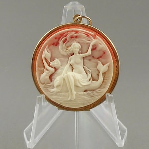 Puede incluir: Colgante de camafeo redondo con marco dorado. El camafeo presenta un relieve tallado de una mujer sentada sobre un delfín, con un fondo rojo y blanco. El colgante se exhibe sobre un soporte de plástico transparente.