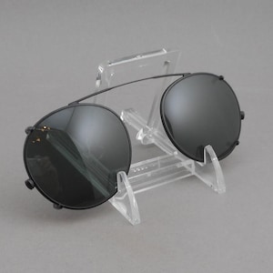 May include: Black round sunglasses with a thin metal frame. The sunglasses have dark lenses and are displayed on a clear plastic stand. The design is a classic, retro style.