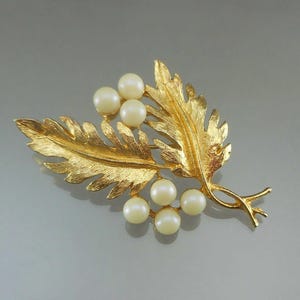 Könnte beinhalten: Goldfarbene Brosche in Form eines Blattes und eines Beerenzweigs. Die Brosche hat strukturierte goldene Blätter und sechs unechte Perlenbeeren. Ein Accessoire im Vintage-Stil.