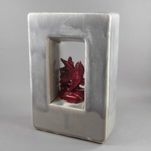 Puede incluir: Un jarrón rectangular de cerámica gris con una abertura cuadrada que revela una escultura roja oscura brillante. La escultura parece ser una forma floral u orgánica estilizada. El jarrón tiene una superficie lisa y reflectante.