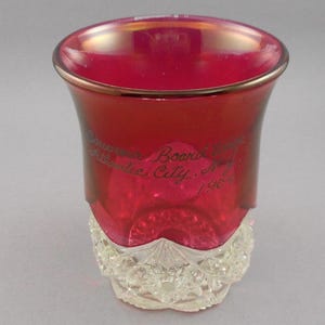 May include: A ruby red glass souvenir shot glass with a clear, patterned base. The glass has a gold rim and features the etched text "Souvenir Board Walk Atlantic City, N.J. 190-".