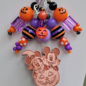 May include: A Halloween-themed charm featuring Mickey and Minnie Mouse dressed in costumes. Mickey is a vampire and Minnie is a witch. They are sitting on a pumpkin with a jack-o'-lantern face. The charm is made of wood and is decorated with orange, purple, and black beads.