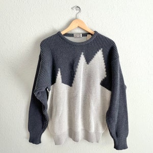 Könnte beinhalten: Ein grauer und weißer Strickpullover mit einem geometrischen Bergdesign. Der Pullover hat einen Rundhalsausschnitt und lange Ärmel.
