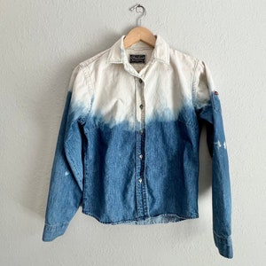May include: A long-sleeved denim shirt with a white ombre effect. The shirt has a button-up front and a patch on the left sleeve.