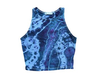 Pode incluir: Um top cropped sem mangas com um padrão tie-dye em tons de azul e roxo. O top tem um decote alto e uma silhueta justa. O tecido parece macio e leve. O top tem uma pequena etiqueta no decote.