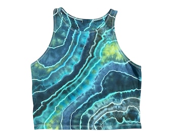 Pode incluir: Um top curto sem mangas com um padrão tie-dye em tons de azul, verde e branco. O tecido tem um efeito marmoreado, com linhas onduladas criando um design único. O top tem um decote alto e é provavelmente feito de um material macio e confortável.