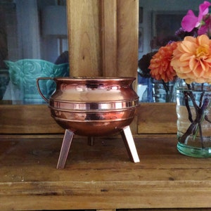 May include: A copper-colored, three-legged cauldron with a handle. The pot has a rounded body with horizontal bands and a polished finish. The background includes a wooden cabinet and a vase of orange flowers.