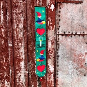 May include: A green fabric banner with a repeating pattern of red hearts, a candle, and a gnome holding a bowl. The banner is hanging on a red wooden door.
