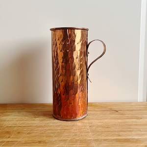 Könnte beinhalten: Ein gehämmerter Kupferkrug mit einem einzigen Henkel. Der Krug hat ein rustikales, gealtertes Aussehen und ist ein wunderschönes Beispiel für handgefertigte Kupferwaren.