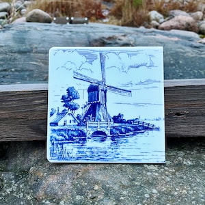 May include: A blue and white ceramic tile depicting a traditional Dutch windmill with a small house and a bridge over a canal. The tile has a blue and white design with a light blue background.