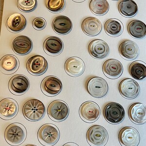 May include: A collection of 36 vintage buttons arranged in a grid on a white background. The buttons are made of various materials, including mother-of-pearl, metal, and plastic. They come in a variety of colors, including white, black, brown, gold, and green. Some of the buttons have intricate designs, while others are plain.