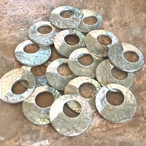 May include: A collection of iridescent, circular shell beads with a central hole. The beads have a cracked, mosaic-like appearance with shades of gray, white, and hints of blue and green. The beads are arranged on a textured, brown surface.