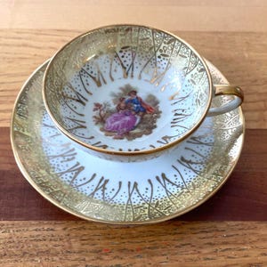 May include: A vintage teacup and saucer set. The white porcelain cup and saucer are adorned with gold filigree and a central image of a couple in period clothing. The cup has a gold handle and rim.