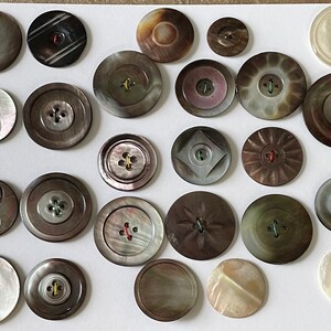 May include: A collection of 20 vintage mother of pearl buttons in various shapes, sizes, and colors. The buttons have a range of designs, including a simple two-hole button, a four-hole button with a starburst design, and a button with a diamond shape.