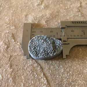 May include: A silver-colored metal object with a textured surface, measured with a ruler. The ruler shows measurements in inches and millimeters. The object is approximately 1 inch in diameter.