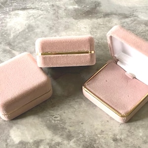 May include: Three pink velvet jewelry boxes. One is open, revealing a white interior. The boxes have gold trim. The closed boxes are rectangular and square. The open box is angled, showing the interior.