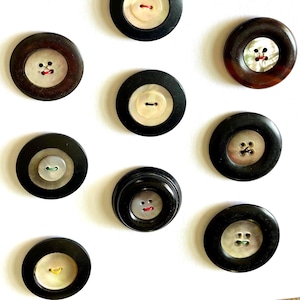 May include: A collection of various vintage buttons. The buttons are primarily black and brown with a variety of off-white, mother-of-pearl, and colored centers. The buttons have two or four holes for sewing.