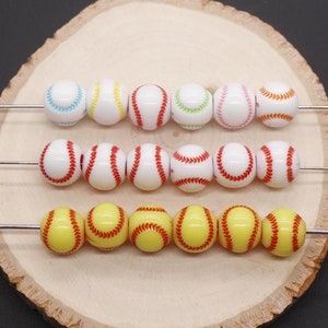 May include: Three rows of baseball-themed beads, each row with a different color scheme. The top row has white beads with blue, yellow, green, pink, and orange stitching. The middle row has white beads with red stitching. The bottom row has yellow beads with red stitching.