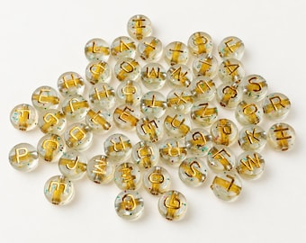 May include: A collection of round, clear acrylic beads featuring embedded gold-colored letters and small, multicolored confetti-like specks throughout the material. Each bead has a central hole, and the letters are printed in a bold, sans-serif font.
