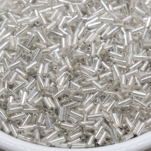 May include: A white bowl filled with silver glass seed beads. The beads are small and cylindrical in shape.