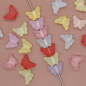 Op de afbeelding: Assortiment van kleurrijke vlinder-vormige kralen in verschillende pasteltinten, waaronder roze, blauw, geel en rood. De kralen zijn geregen aan een zilveren draad.