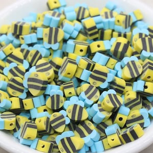 May include: A bowl filled with yellow and black bee-shaped beads with blue accents. The beads are made of polymer clay and are perfect for jewelry making or crafting.