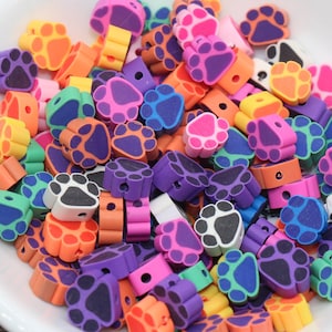 Puede incluir: Un bol lleno de cuentas de plástico con forma de huella de pata de colores. Las cuentas están disponibles en una variedad de colores, incluyendo morado, azul, verde, naranja, rosa y negro.