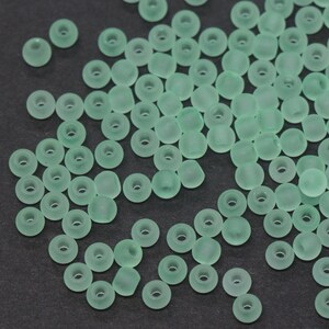 May include: A collection of small, round, seafoam green beads with a matte finish. Each bead has a hole running through the center, and they are scattered across a dark surface. These beads are suitable for jewelry making and crafts.