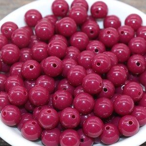 May include: A white bowl filled with approximately 50 round, deep burgundy-colored beads. The beads are made of a glossy, smooth material.