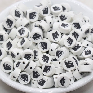 May include: A white bowl filled with white round beads with black graduation cap designs.