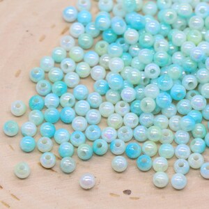 May include: A collection of iridescent, round beads in shades of light blue, turquoise, and white. The beads are scattered on a light brown wooden surface, creating a visually appealing display. These beads are ideal for jewelry making and craft projects.