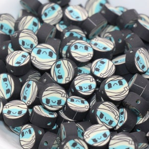 May include: A bowl filled with black and white mummy-themed polymer clay beads. The beads have a light blue face with a black mouth and eyes.