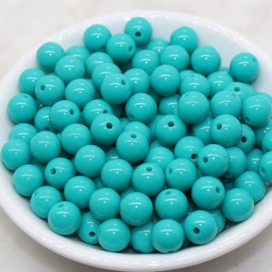 May include: A white bowl filled with turquoise round beads. Each bead has a small hole. The beads are a solid, glossy color. The bowl is sitting on a white surface. These beads are suitable for jewelry making and crafts.